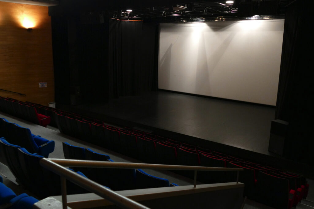 Image de la salle de cinéma montrant les sièges bleus et rouges, la scène et l'écran de cinéma prise depuis le haut de la salle.